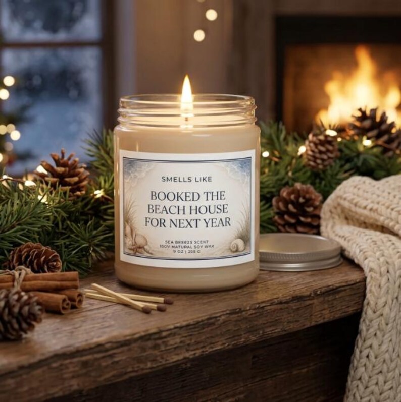 Lit soy candle labeled Smells Like Booked the Beach House For Next Year, styled with pine, pinecones and a cozy knit blanket by a fireplace
