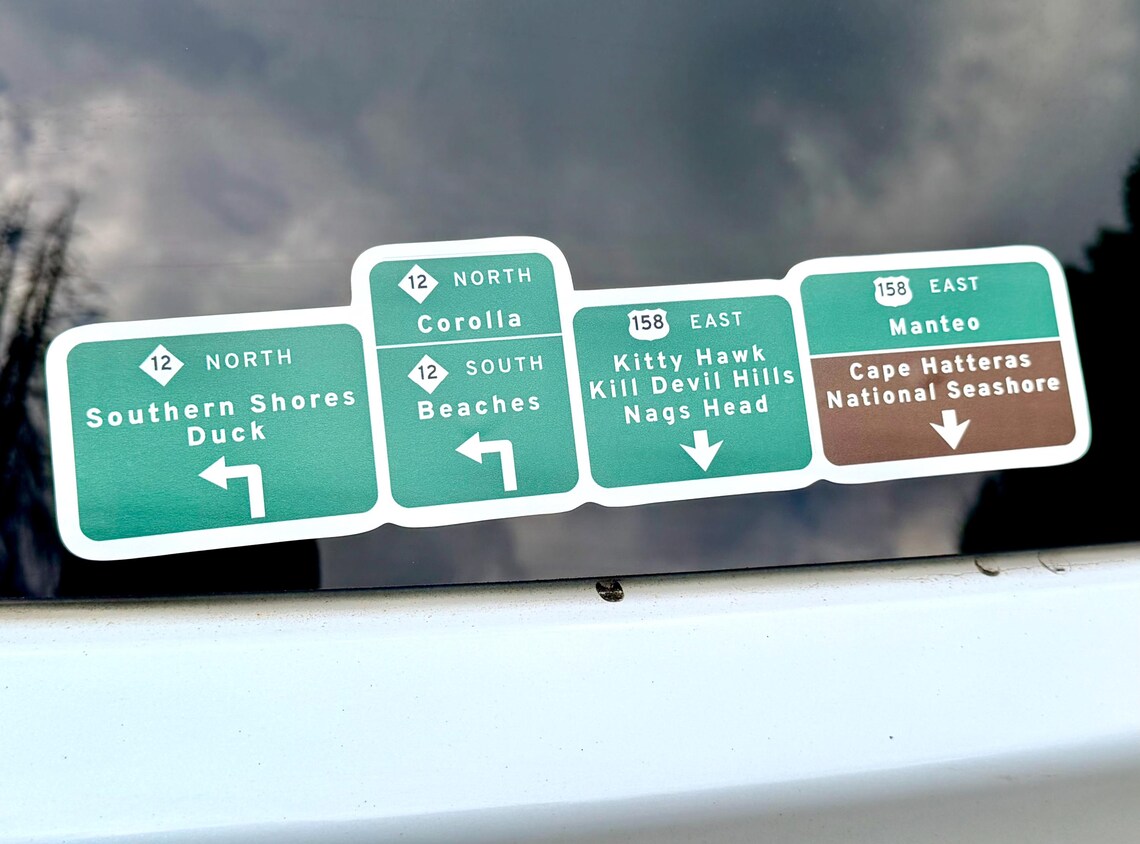 Green Outer Banks highway sign vinyl decal on a car window, listing Southern Shores, Duck, Corolla, Kitty Hawk, Kill Devil Hills, Nags Head, Manteo and Cape Hatteras National Seashore